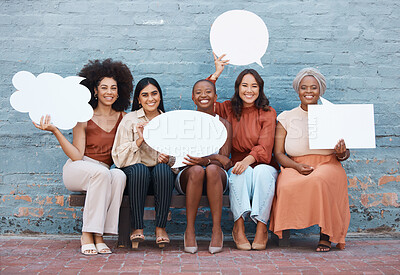 Group of five diverse young cheerful businesswomen sitting on a bench against a wall outside in the city and holding speech bubbles. Businesspeople smiling holding contact detail boards outdoors together