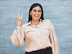 One young business woman of indian ethnicity standing outside against a grey wall and gesturing the peace sign with her hand. Happy and confident mixed race executive looking positive and successful