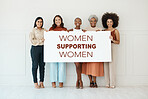 Group of five diverse young businesswomen standing against a wall in an office and holding a sign in support and unity. Happy colleagues holding a sign with a message while standing in a row together at work