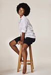 Studio portrait of a young stunning African American woman with a beautiful afro.Confident black female model showing her smooth complexion while sitting on a chair and posing against a grey background