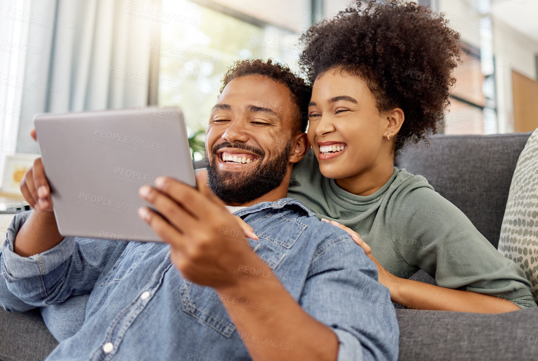 Buy stock photo Mixed race couple smiling while using a digital tablet together at home. Joyful hispanic boyfriend and girlfriend laughing while relaxing and using social media on a digital tablet in the lounge at home