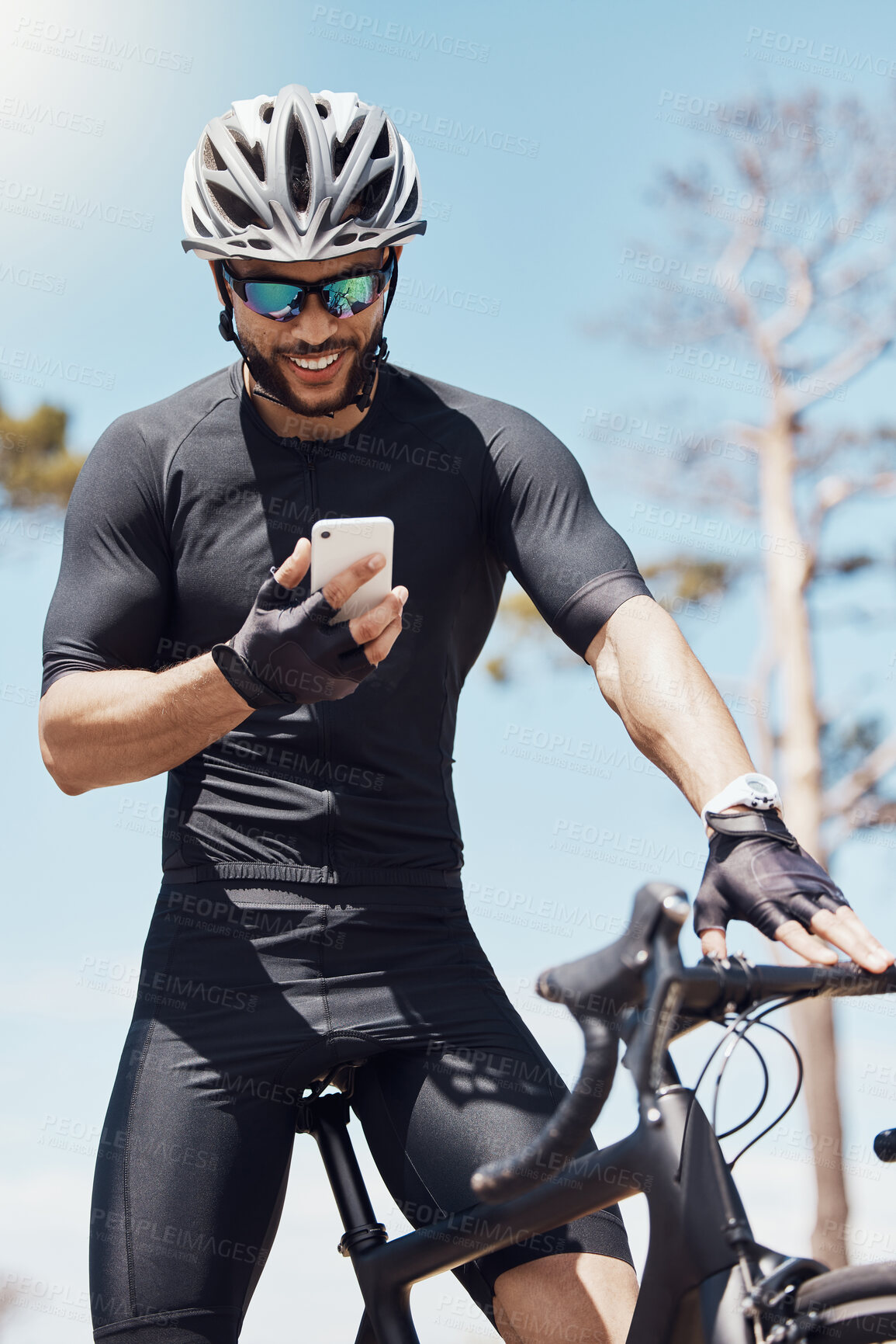 Buy stock photo Smiling young male cyclist wearing helmet and glasses while using his smartphone and standing with his bike. Fit sportsman using app or sending a message on mobile phone while cycling outdoors