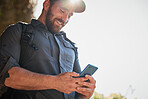 Happy caucasian man using smartphone while carrying a backpack and hiking alone in the mountains on a sunny day. Smiling fit and active man using mobile app or messaging while in a nature environment  