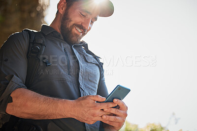 Happy caucasian man using smartphone while carrying a backpack and hiking alone in the mountains on a sunny day. Smiling fit and active man using mobile app or messaging while in a nature environment