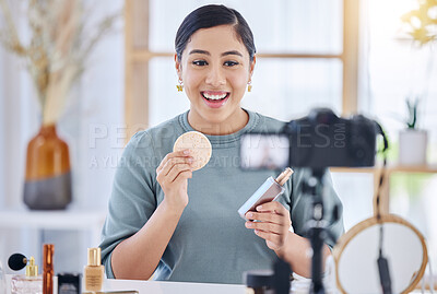 Smiling young beauty vlogger using her camera to make an online video applying cosmetic makeup to her face. Happy young woman applying makeup to her holding a product and talking to a camera at home