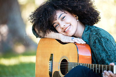 Smiling young musician resting on her acoustic guitar while playing and enjoying a day in the park. Happy hispanic woman with curly afro hair playing music on her guitar in the garden outside