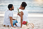 African American couple spending a day at the sea together. Content girlfriend smelling a rose with her boyfriend while sitting on the beach. Caring husband and wife bonding on the seashore