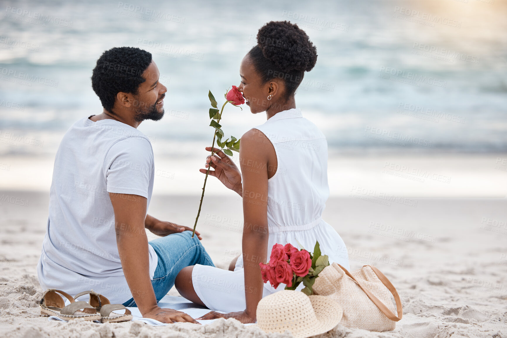 Buy stock photo African American couple spending a day at the sea together. Content girlfriend smelling a rose with her boyfriend while sitting on the beach. Caring husband and wife bonding on the seashore