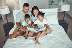 Young mixed race parents mom and dad reading a book to little daughter and son while bonding and spending time together at home in the morning. Two children enjoying fairy tale story while sitting with their parents in their bedroom