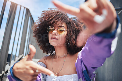Young stylish mixed race woman with curly natural afro hair wearing trendy sunglasses and bright purple vintage jacket making a finger frame gesture. Fashionable and confident woman relaxing outdoors