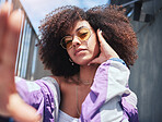 Portrait of young trendy beautiful mixed race woman with an afro taking selfies and posing alone outside. Hispanic woman with curly hair wearing sunglasses. Fashionable African American woman in city