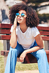 Young stylish mixed race woman with curly afro hair wearing trendy sunglasses and relaxing on a bench at the park. One female only looking carefree, cool and confident while enjoying sunny day outside
