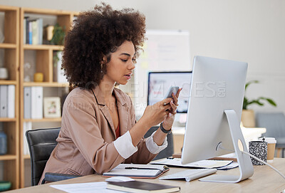 One young busy African American woman using a desktop and cellphone at a desk in her office job. Black woman with an afro checking social media at her job. Mixed race woman focused on texting