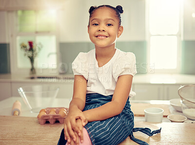 Portrait of a cute young mixed race girl sitting on the kitchen counter smiling and wearing an apron looking thoughtful. Little innocent hispanic girl smiling and sitting alone after baking in the kitchen at home. Baking is one of her hobbies and she dayd