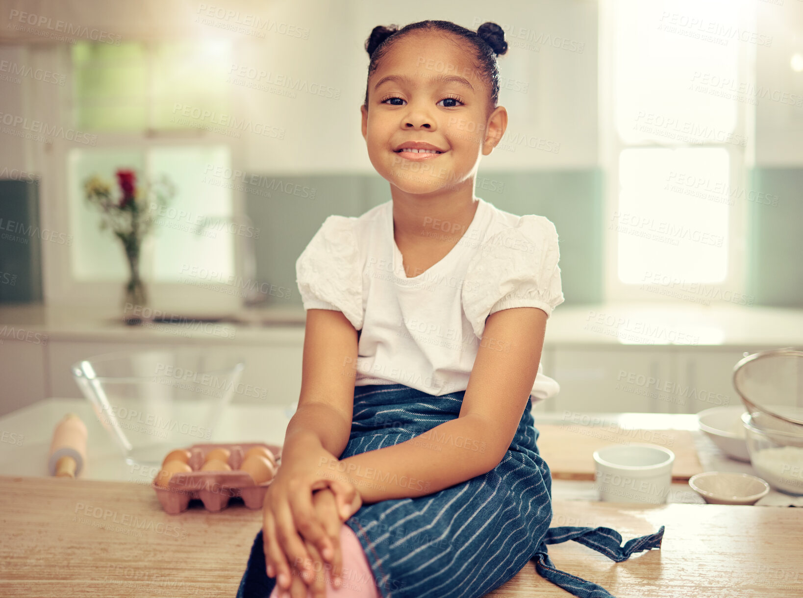 Buy stock photo Portrait of a cute young mixed race girl sitting on the kitchen counter smiling and wearing an apron looking thoughtful. Little innocent hispanic girl smiling and sitting alone after baking in the kitchen at home. Baking is one of her hobbies and she daydreams of it everyday