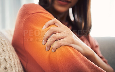 Closeup shot of a mixed race woman in casual wear holding her arm while suffering pain and sitting on her sofa in the lounge at home. One unrecognizable female struggling with an injury and arthritis