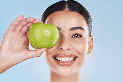Buy stock photo Apple, beauty and skin of a woman with a smile holding a fruit on her face against a blue studio background. Portrait of a happy female model in skincare or dental health smiling with juicy teeth
