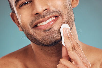 Buy stock photo Closeup of one smiling mixed race man wiping a round cotton swab on his face while grooming against a blue studio background. Handsome guy cleaning and exfoliating his face for a healthy complexion and clear skin