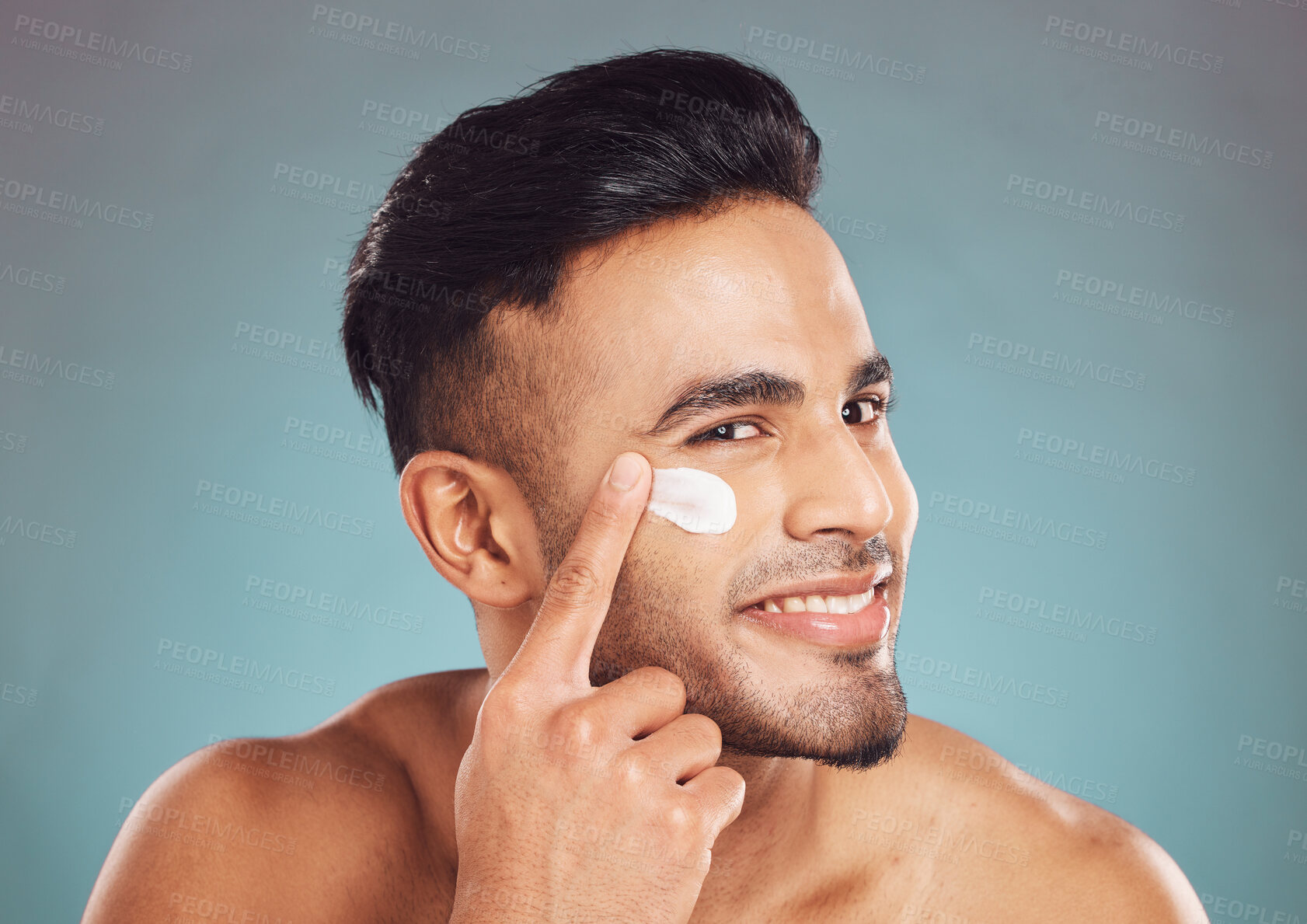 Buy stock photo Portrait of one smiling young indian man applying moisturiser lotion to his face while grooming against a blue studio background. Handsome guy using sunscreen with spf for uv protection. Rubbing facial cream on cheek for healthy complexion and clear skin
