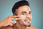 Portrait of one smiling young indian man applying moisturiser lotion to his face while grooming against a blue studio background. Handsome guy using sunscreen with spf for uv protection. Rubbing facial cream on cheek for healthy complexion and clear skin