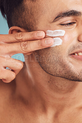 Buy stock photo Closeup of one young indian man applying moisturiser lotion to his face while grooming against a blue studio background. Handsome guy using sunscreen with spf for uv protection. Rubbing facial cream on cheek for healthy complexion and clear skin