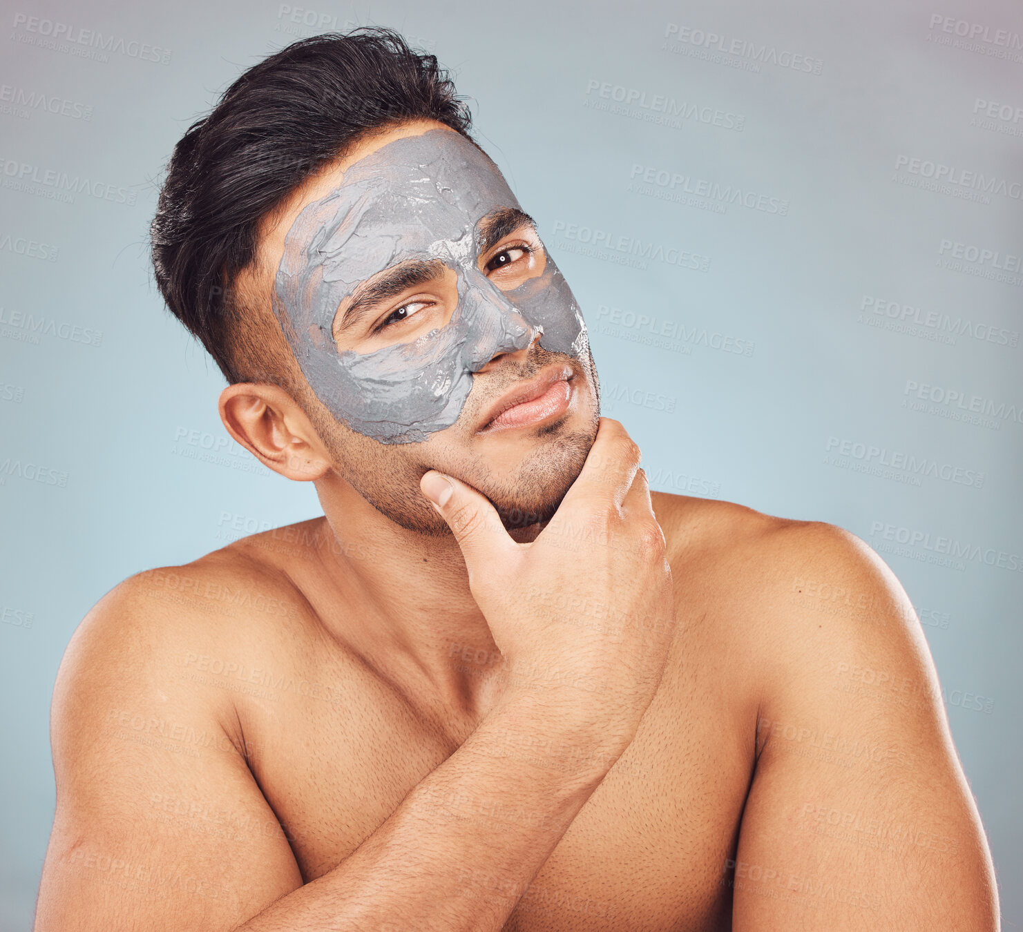 Buy stock photo Portrait of one handsome young indian man applying an anti aging facial mask against a blue studio background. Mixed race guy wearing a moisturising clay or charcoal cream product on his face to get rid of blackheads for healthy, smooth and soft skin