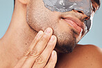 Closeup of one mixed race man touching his beard and chin while applying a detoxifying facial mask against a blue studio background. Guy wearing a moisturising clay or charcoal cream product on his face for healthy, smooth soft skin