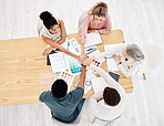 Group of five diverse businesspeople having a meeting at a table in an office from above. Happy business professionals talking while stacking their hands in support at work