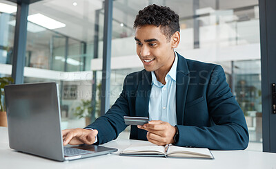 Young happy businessman holding and using a credit card and typing on a laptop alone at work. One hispanic male businessperson smiling while making an online payment with his debit card and laptop