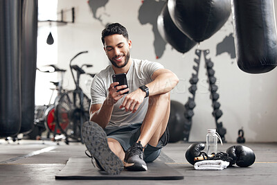 Buy stock photo One fit young hispanic man using a cellphone while taking a break from exercise in a gym. Happy mixed race guy texting and browsing fitness apps online while checking social media during a rest from training workout