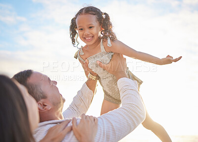 Buy stock photo Cute mixed race girl being lifted into the air by her mixed race father while her mother watches. Adorable daughter and her parents spending time together by the ocean at sunset. Family bonding