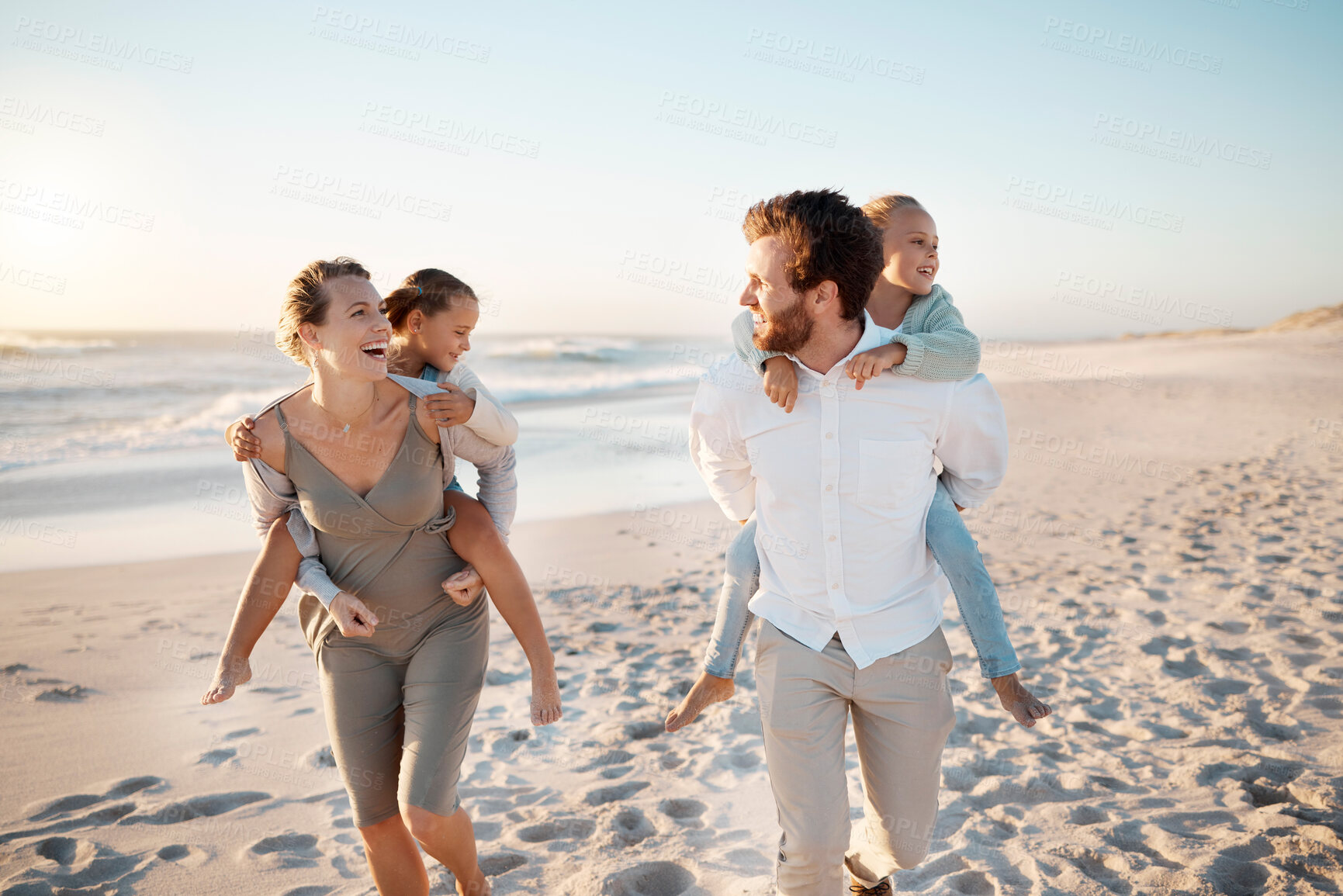Buy stock photo Playful parents enjoying a holiday with their children. Little girls getting piggyback rides from their parents. Carefree family on holiday by the beach together. Family bonding by the ocean