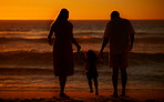 Rearview cute mixed race girl standing hand in hand with her mom and dad in the sea at the beach. A young couple and their daughter holding hands while standing in the water and looking at a sunset