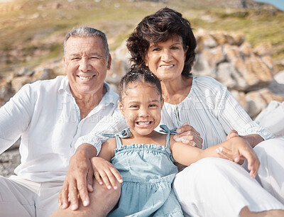 Buy stock photo Smiling mixed race grandparents sitting with granddaughter on a beach. Adorable, happy, hispanic girl bonding with grandmother and grandfather outside on weekend. Seniors and child enjoying free time