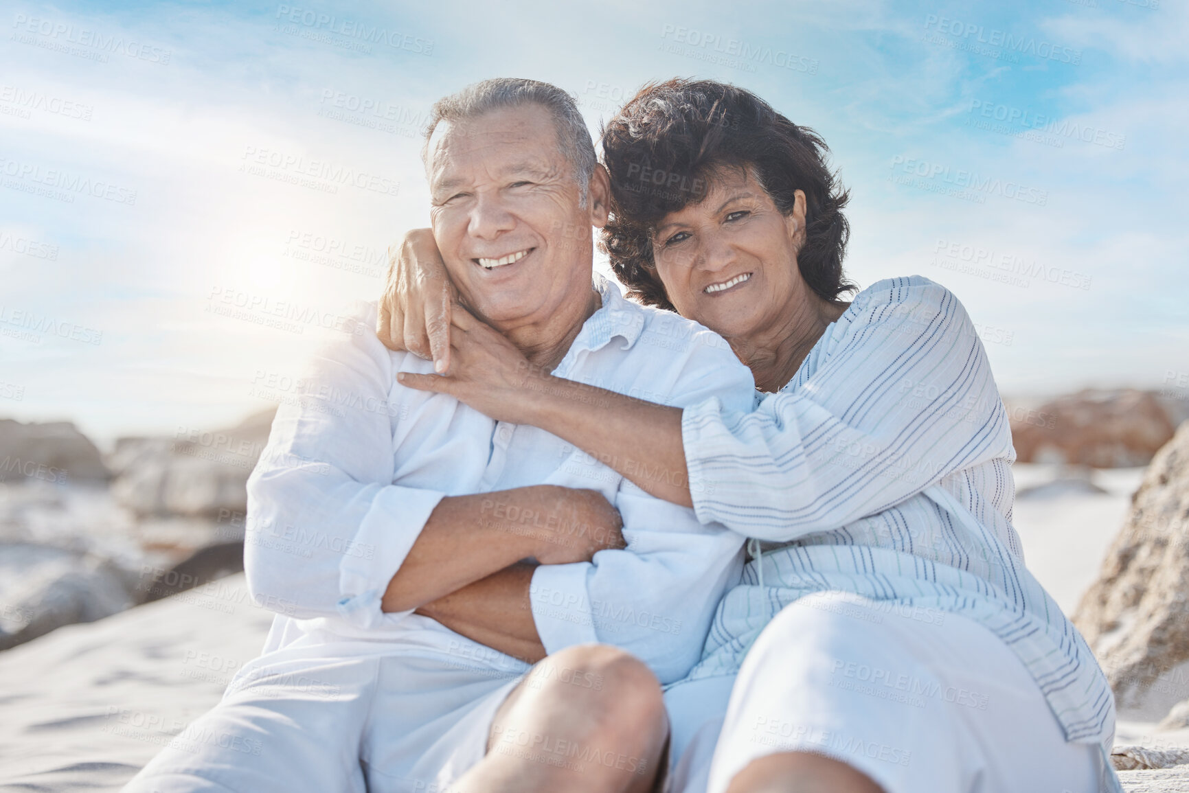 Buy stock photo Portrait of a senior mixed race couple sitting together on the beach embracing one another and smiling on a day out at the beach. Hispanic husband and wife looking happy and showing affection while having a romantic day on the beach