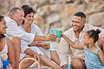 Closeup of a mixed race family having a picnic on the beach and smiling  while having some food with snacks. Happy family bonding on a day out at the beach