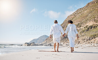 Buy stock photo Rear view of a Mixed race senior couple taking a  romantic walk on the beach and holding hands on a sunny summer day outdoors