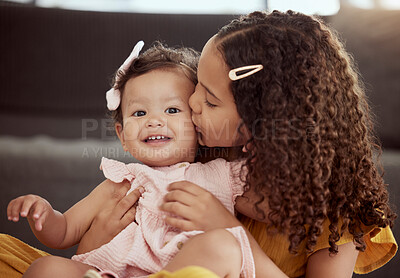 Adorable little mixed race child kissing baby sister on the cheek at home. Two small cute hispanic girls sitting together and bonding in living room. Affectionate sibling with curly hair and toddler