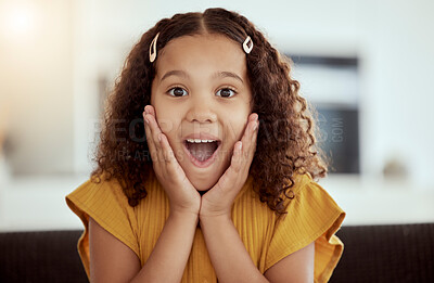 Adorable little mixed race child touching face with her hands in surprise at home. One small cute hispanic girl sitting alone on a living room sofa and looking shocked. Excited kid with curly hair