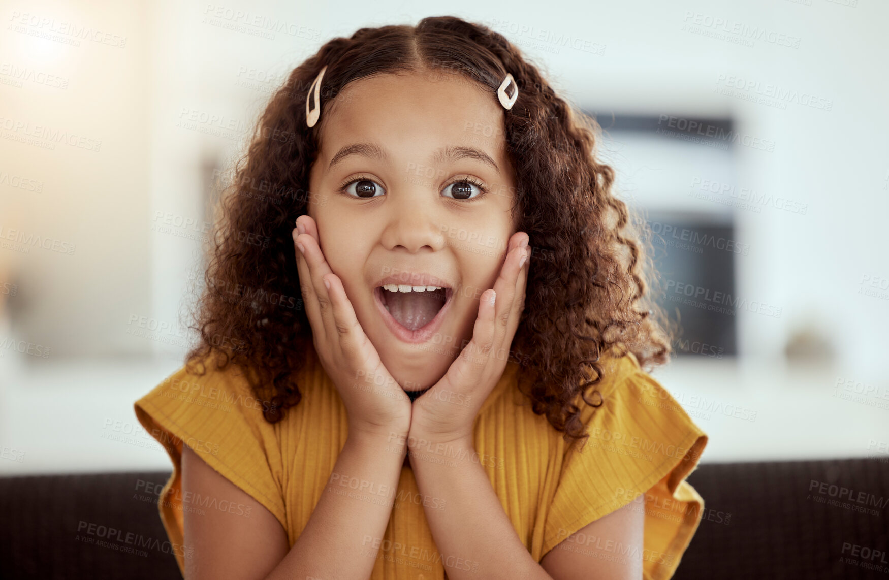 Buy stock photo Adorable little mixed race child touching face with her hands in surprise at home. One small cute hispanic girl sitting alone on a living room sofa and looking shocked. Excited kid with curly hair