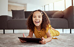 Portrait of adorable little mixed race child using digital tablet in home living room. One small cute hispanic girl lying alone on lounge floor, playing game on technology. Happy kid with curly hair