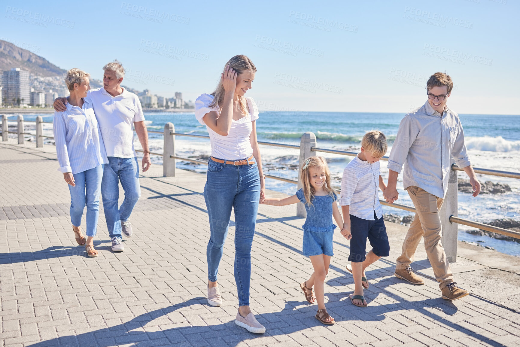 Buy stock photo Loving caucasian parents holding hands with their little daughter and son while their grandparents follow in the background. Happy multi-generation family walking along seaside promenade on a sunny day
