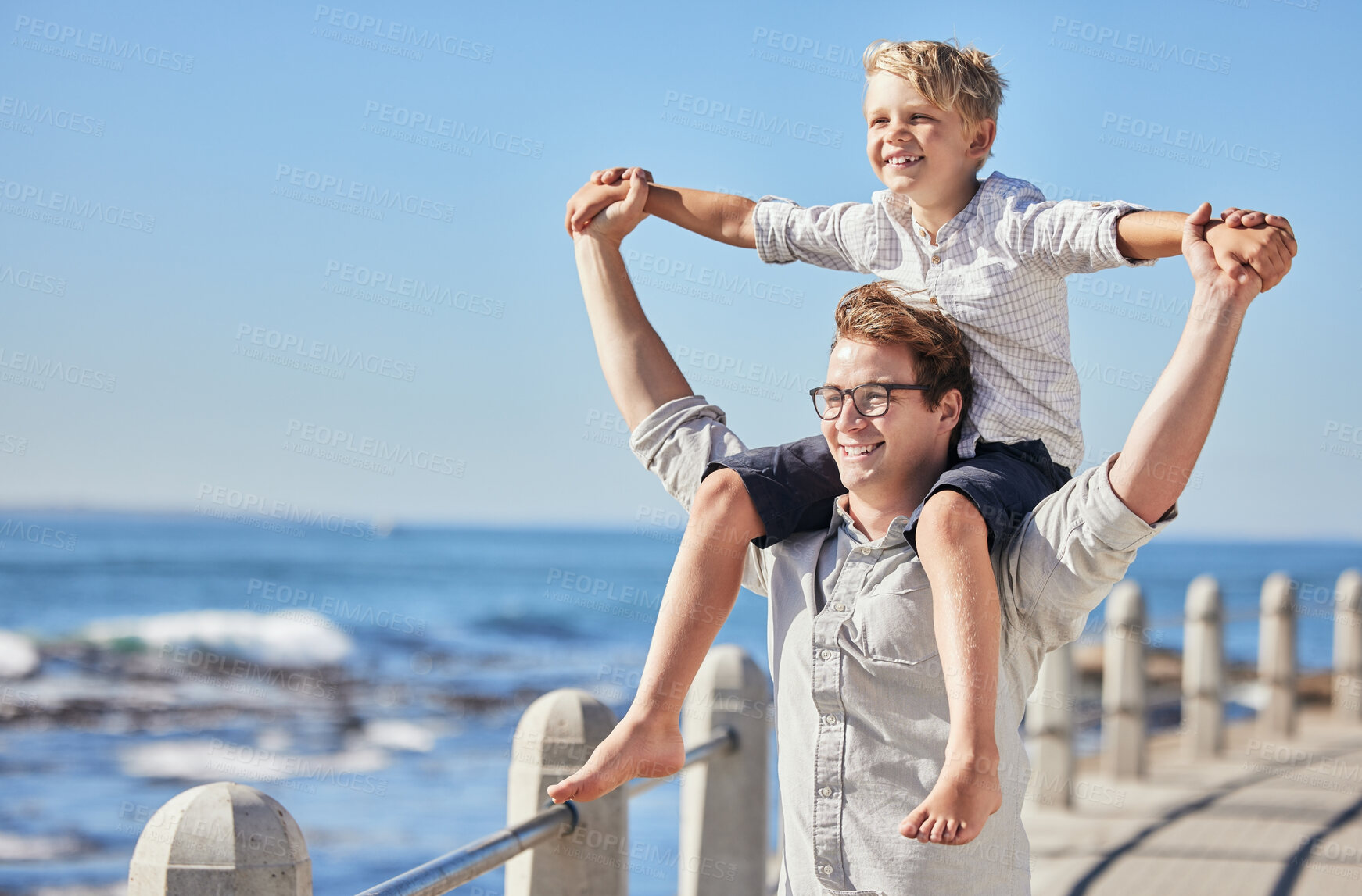 Buy stock photo Excited little boy holding hands with his father while sitting on his shoulders as they walk along the promenade. Happy father and son having fun and spending time together by the sea while on vacation