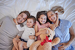 Portrait of a happy caucasian family with two children relaxing and lying together on a bed at home, from above. Little brother and sister holding stuffed animals and touching mom and dads face
