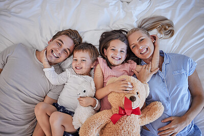 Portrait of a happy caucasian family with two children relaxing and lying together on a bed at home, from above. Little brother and sister holding stuffed animals and touching mom and dads face