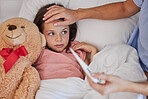 Sick little girl in bed with her teddybear while her mother uses a thermometer to check her temperature. Young concerned single parent sitting with sick child while and feeling her forehead . Small child feeling ill while her mother checks fever