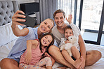 Happy young caucasian family sitting together on bed and taking a selfie or doing video call together in the morning. Carefree mother holding mobile phone while recording video of special moment with her family