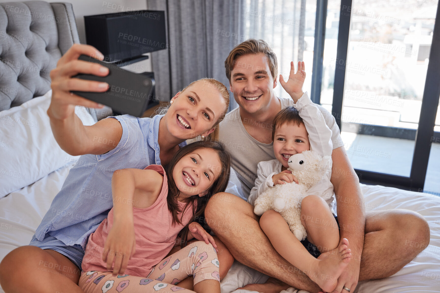 Buy stock photo Happy young caucasian family sitting together on bed and taking a selfie or doing video call together in the morning. Carefree mother holding mobile phone while recording video of special moment with her family