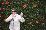 Funny old woman dancing listening to music on smartphone wearing earphones smiling enjoying fun celebrating retirement in garden with flower wall