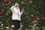 Funny old woman dancing listening to music on smartphone wearing earphones smiling enjoying fun celebrating retirement in garden with flower wall
